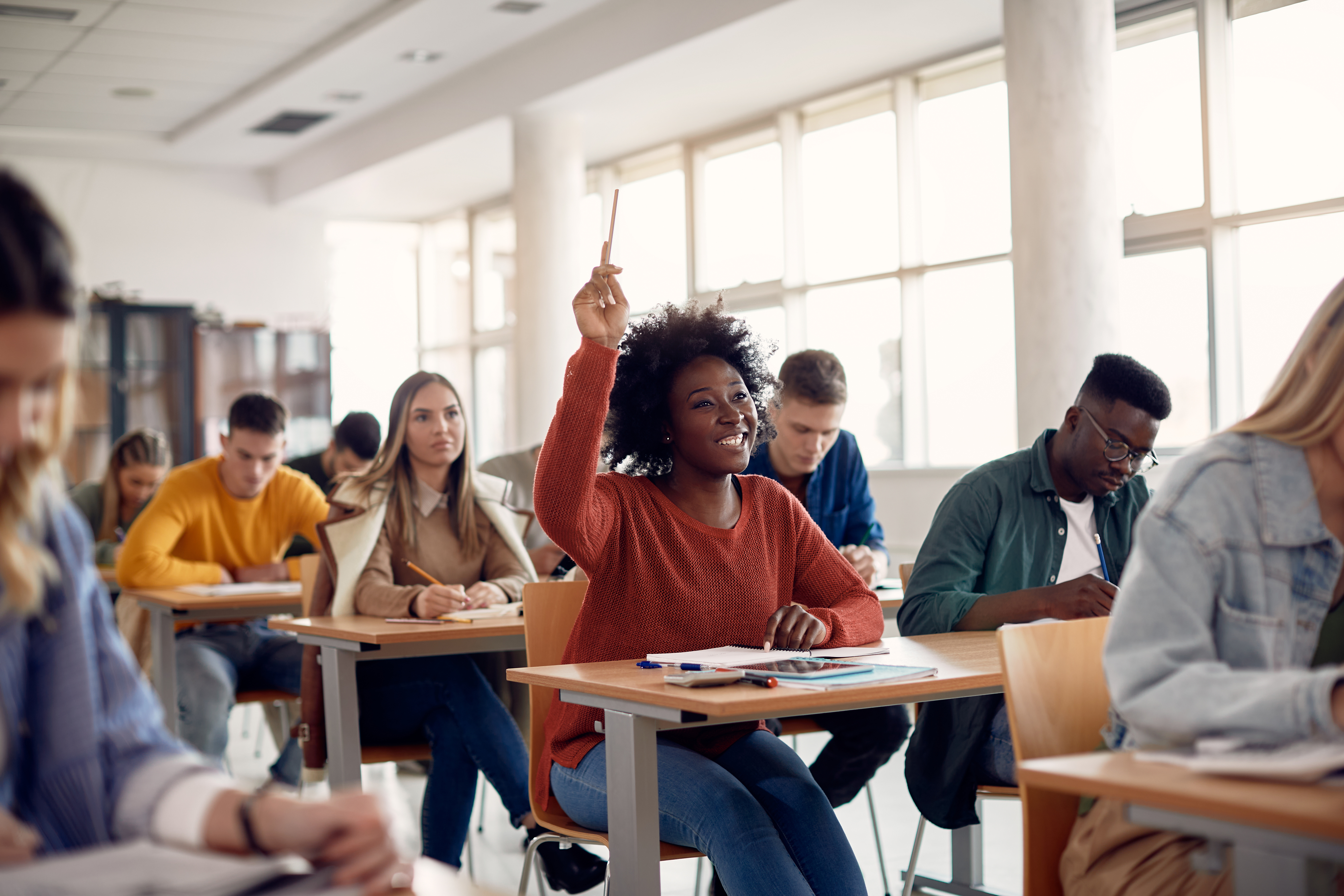 Image shows a classroom of adult students at their desks. In the center of the image is a young woman smiling with her hand raised and a pen in her hand, waiting to be called on by the teacher.