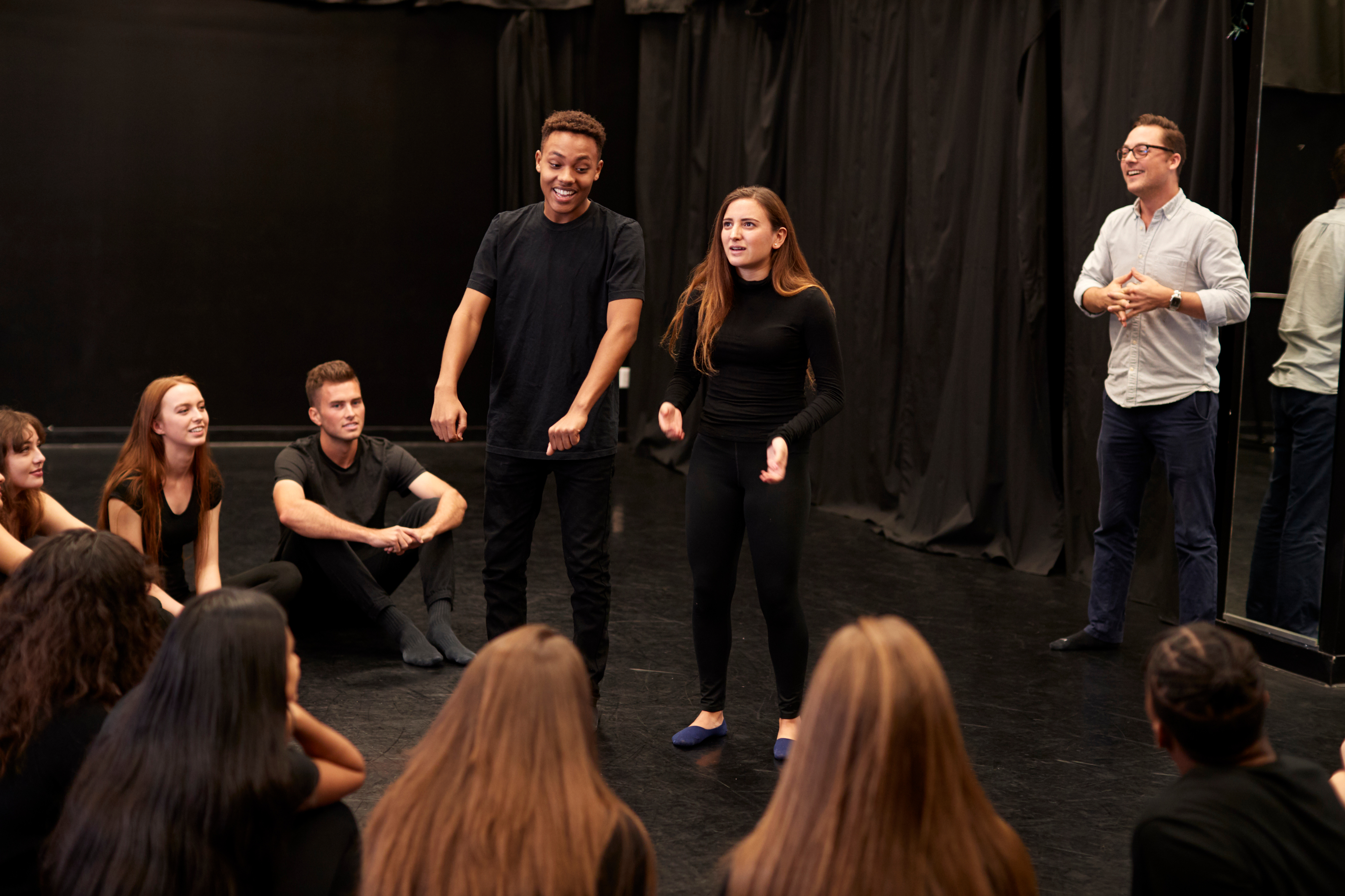 Image shows a high school acting class in a room with a black background. The students are all dressed in black and most are sitting in a circle on the floor with two standing in the middle doing improvisation while the other students and teacher watch.