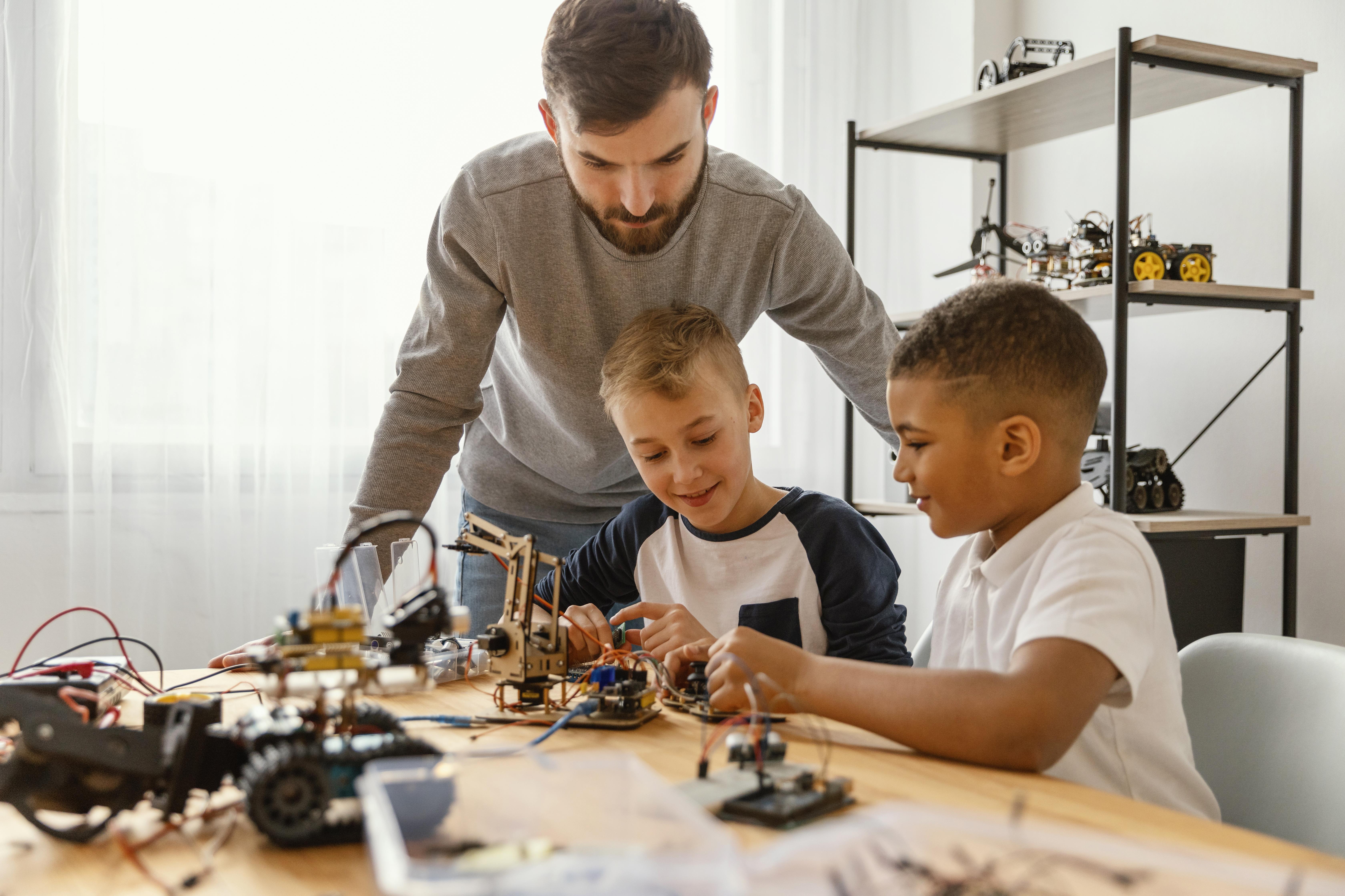 A male teacher is standing over a table and watching two boys who are sitting down and building robots.