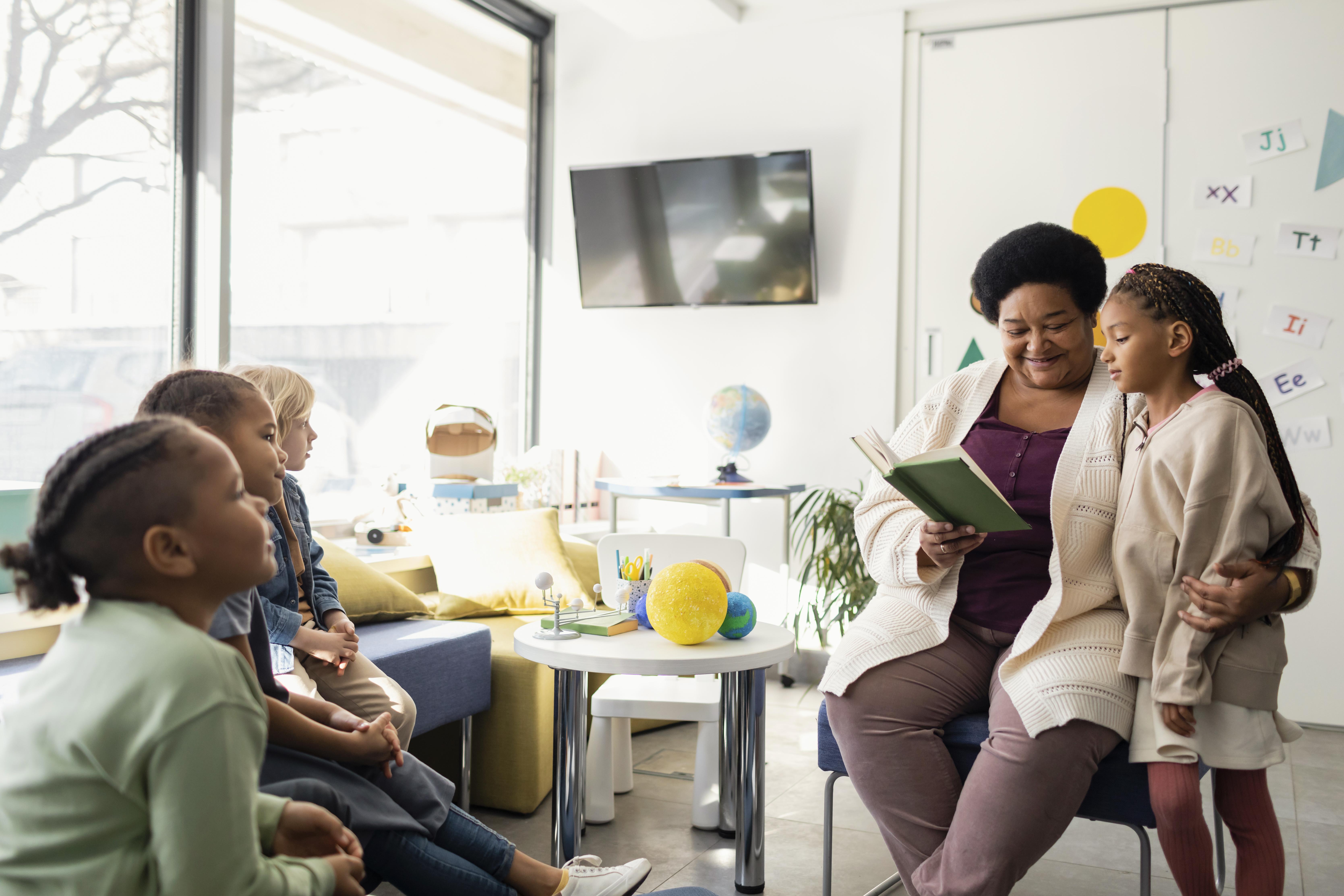 A female teacher is sitting on a chair, helping one of her students read a book aloud to the rest of the class. Image by Freepik