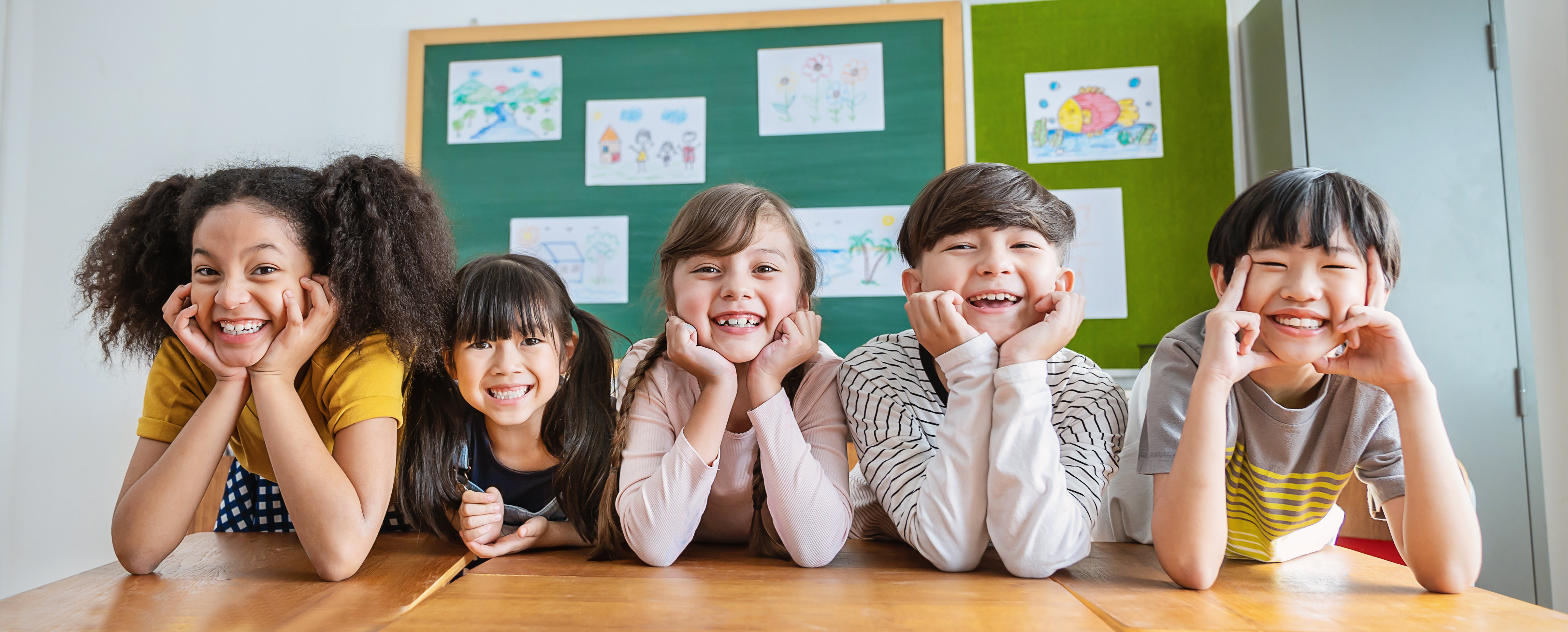 The image shows five children sitting with their elbows propped up on a desk, smiling at the camera.