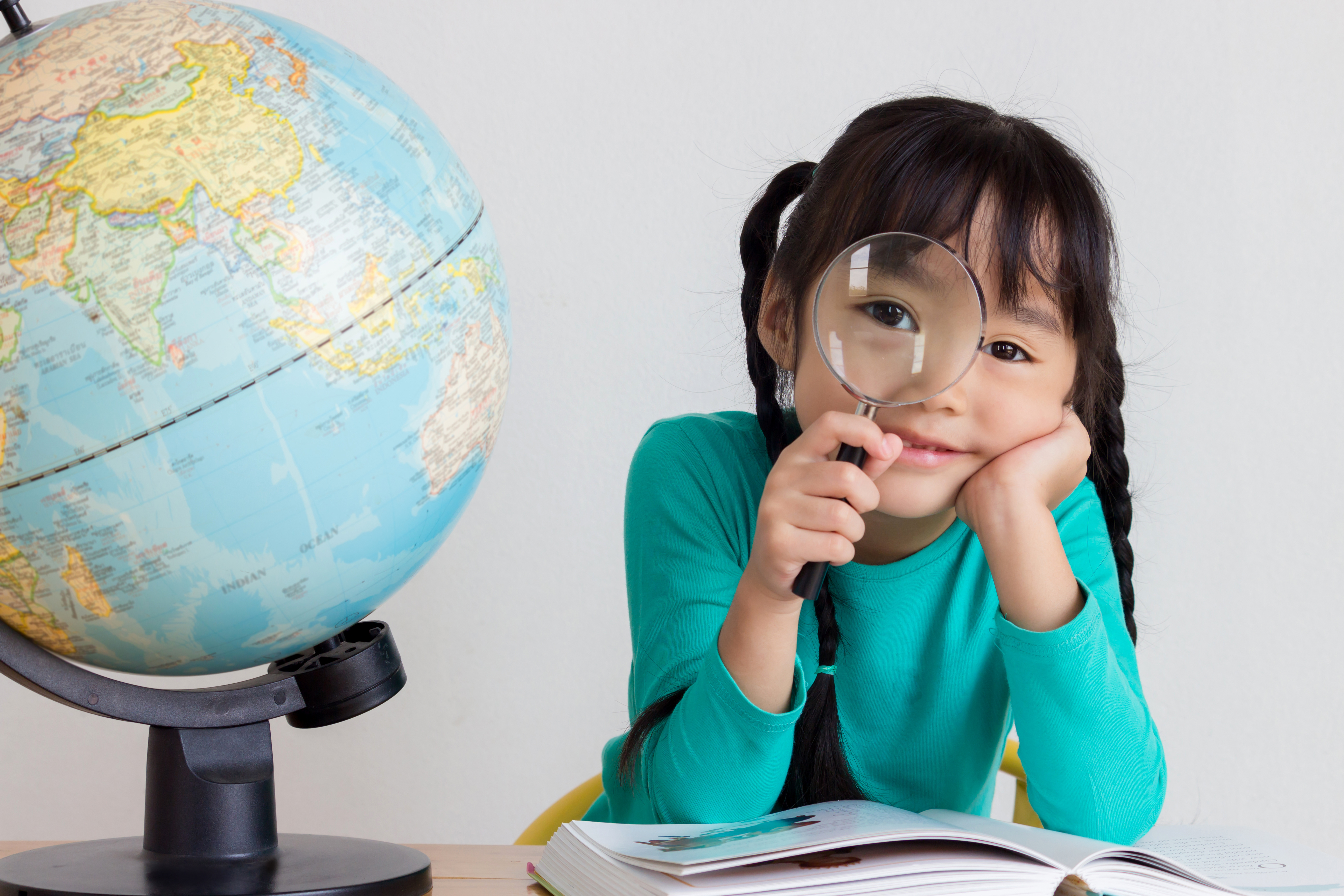 A girl with an open book in front of her is looking at the viewer and holding a magnifying glass to her eye. Next to her on the table is a globe.