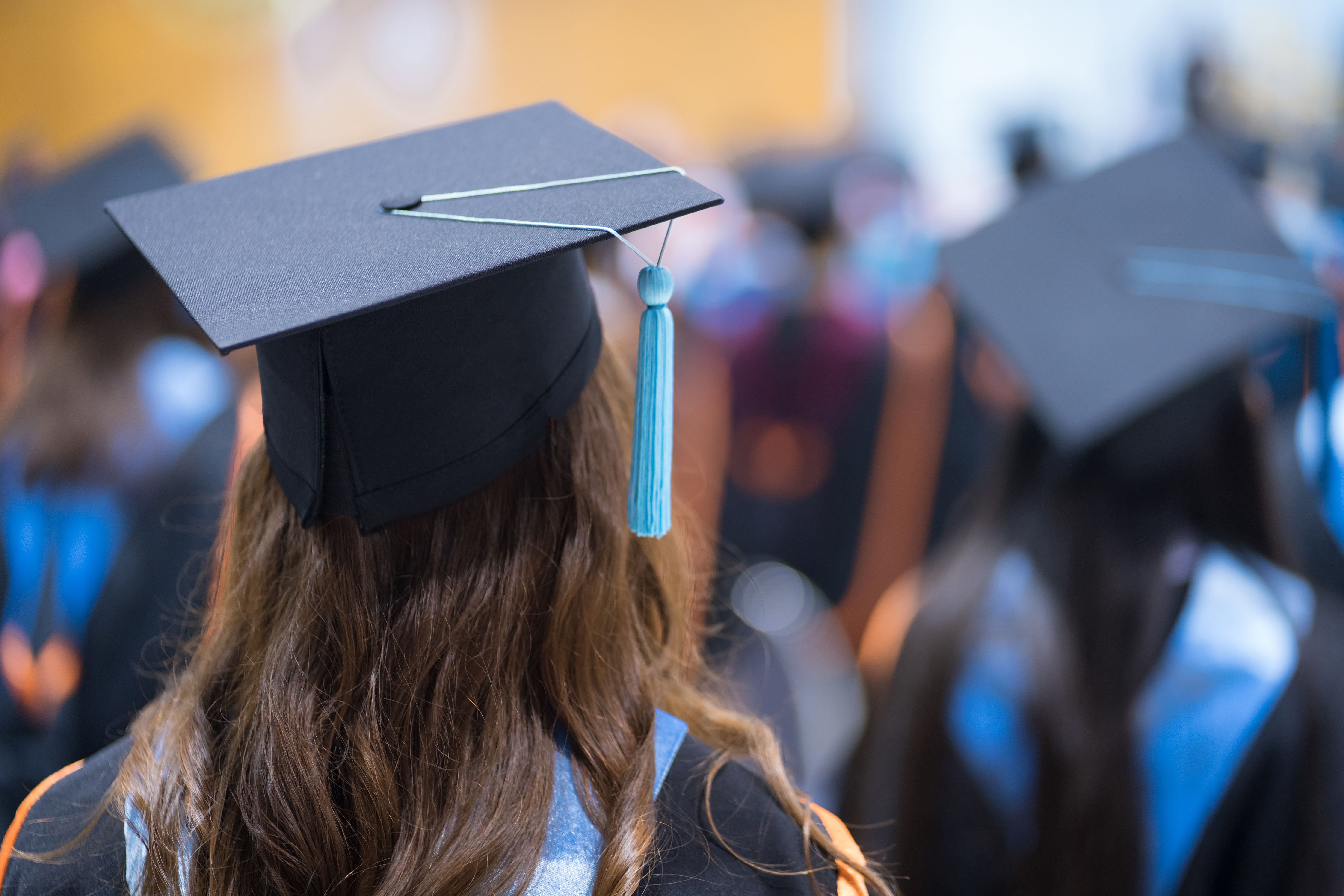 In the foreground, the head and shoulders of a female student with long hair, wearing a graduation cap and gown. In the background, also with their backs to the viewer, are more students dressed in graduation caps and gowns.