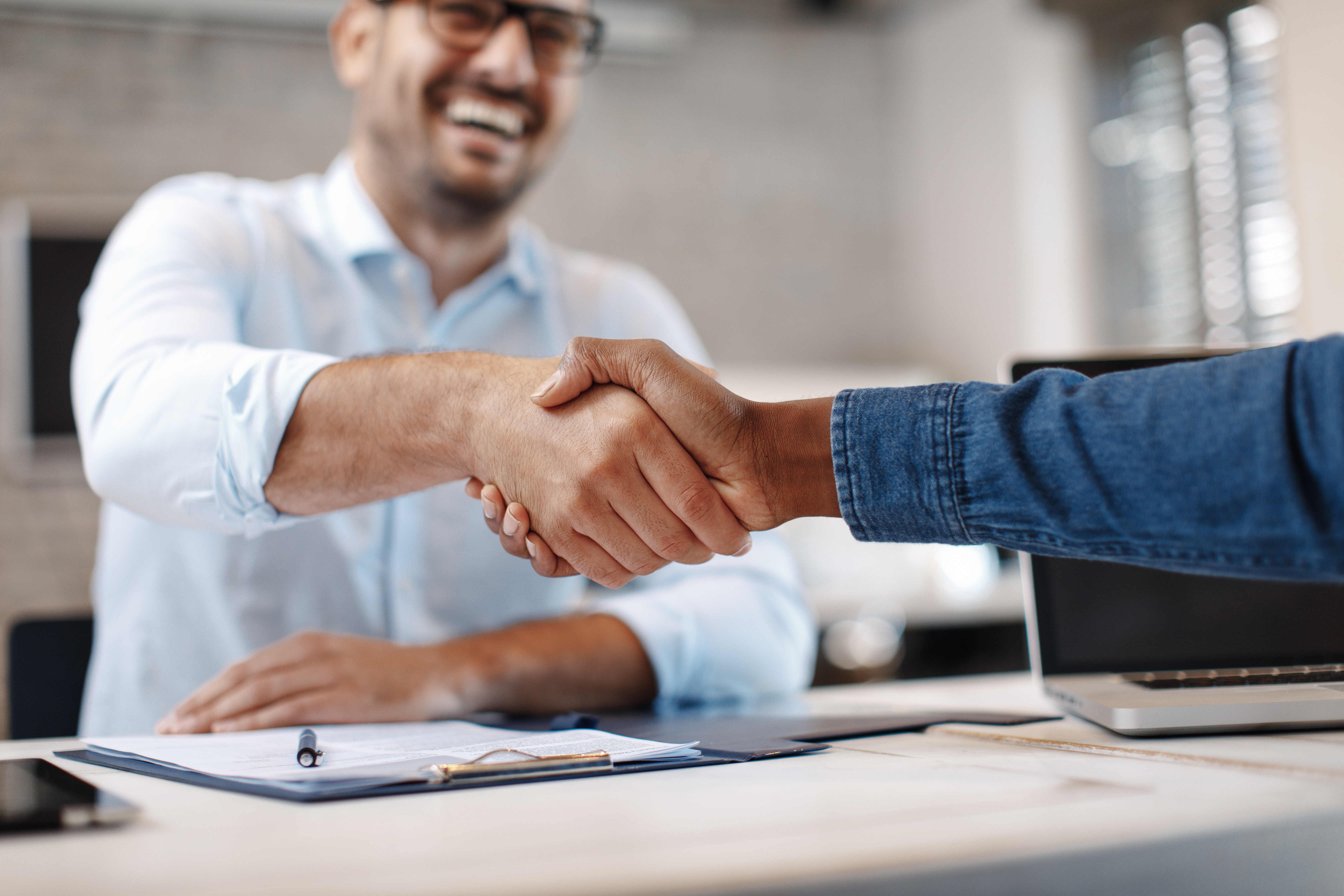 A handshake between a smiling man behind a desk and someone who is out of frame except for their arm.