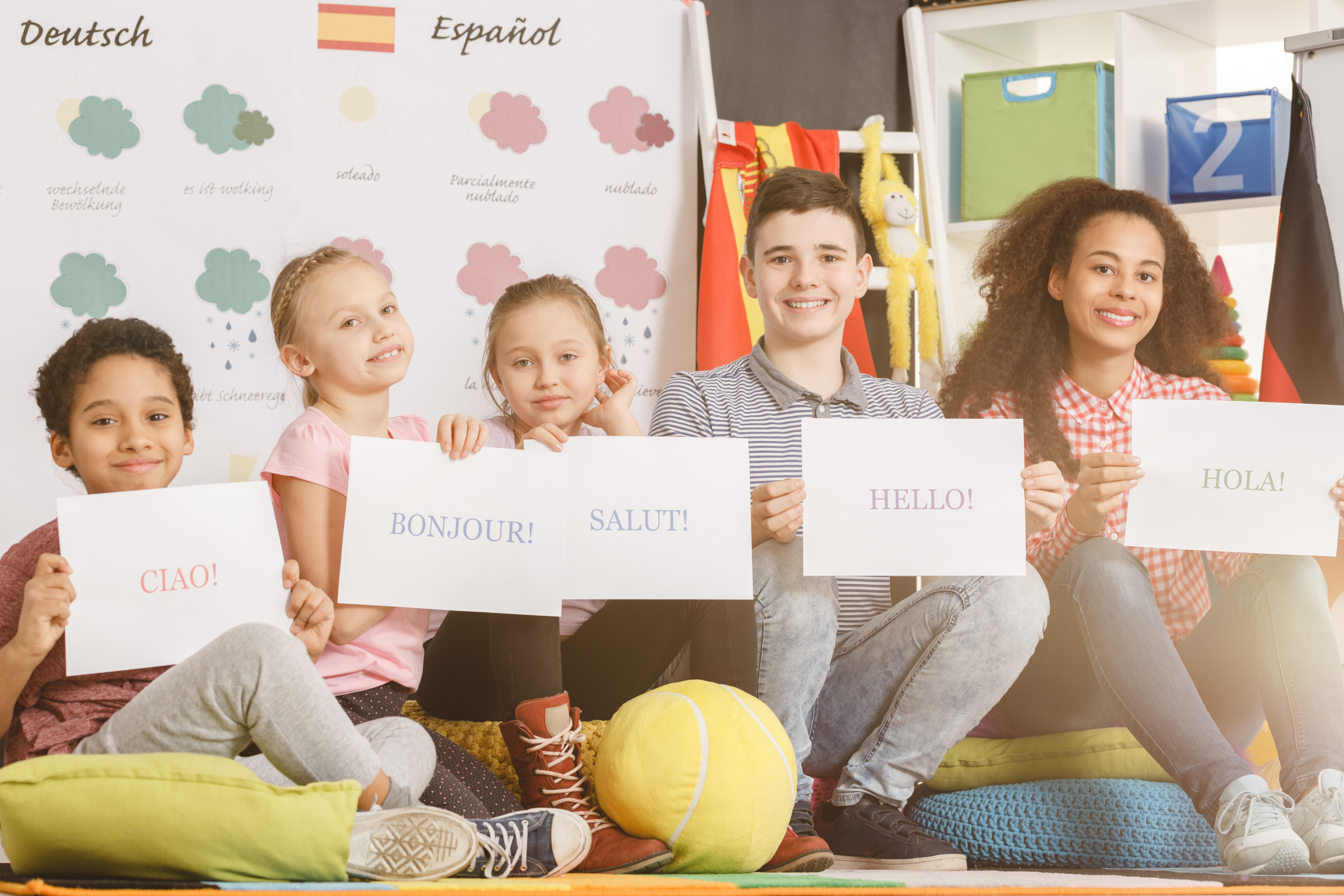 Five children are sitting on the ground holding signs each with a greeting in a different language.  In the background is a classroom with posters of Dutch and Spanish vocabulary.