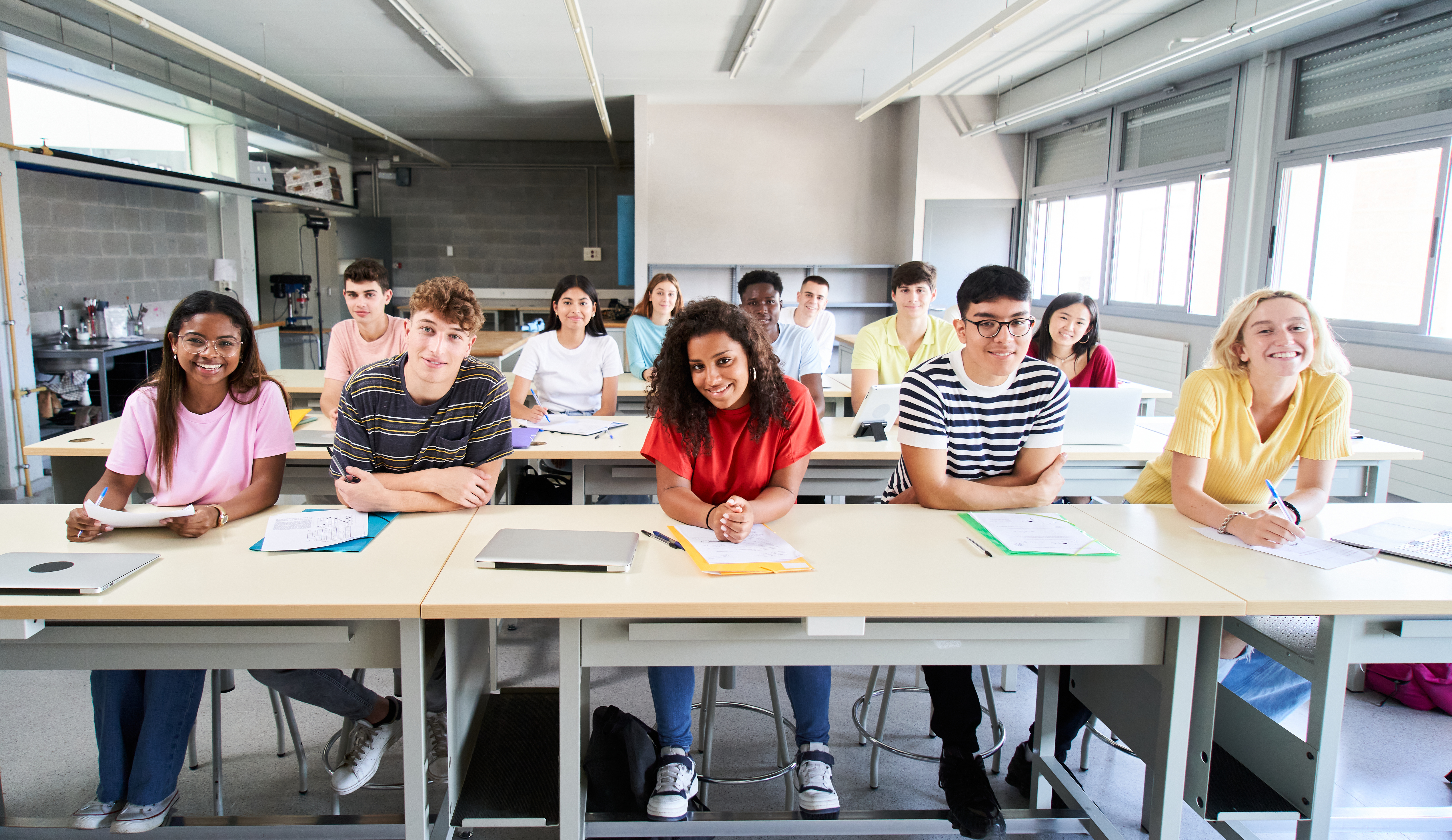 High school students are sitting at their desks in their classroom, smiling and looking at the front of the room where the viewer is.