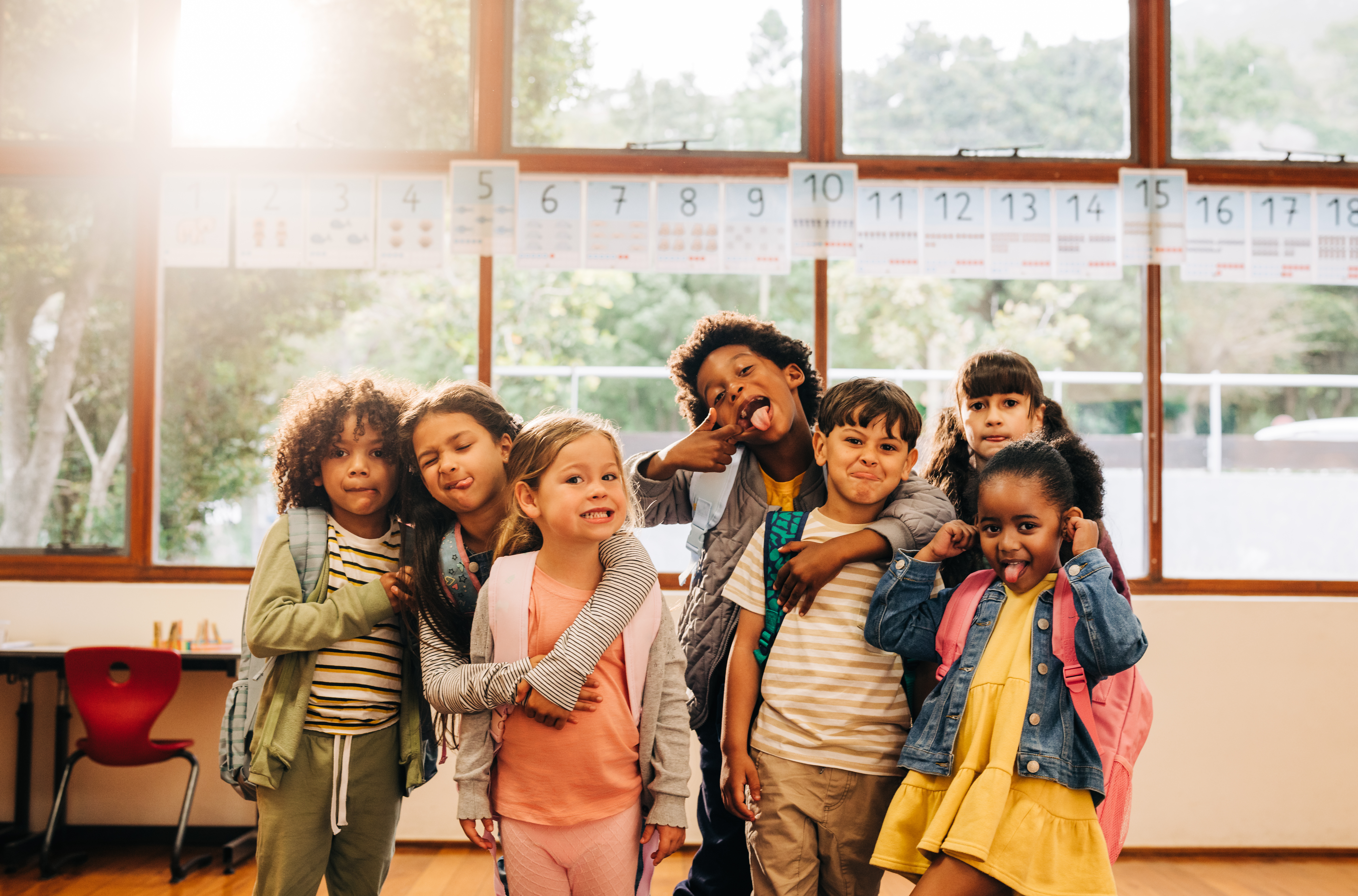 Image shows 7 elementary-aged students wearing backpacks in a classroom looking at the camera and making silly faces. Some of the students have their arms around each other.