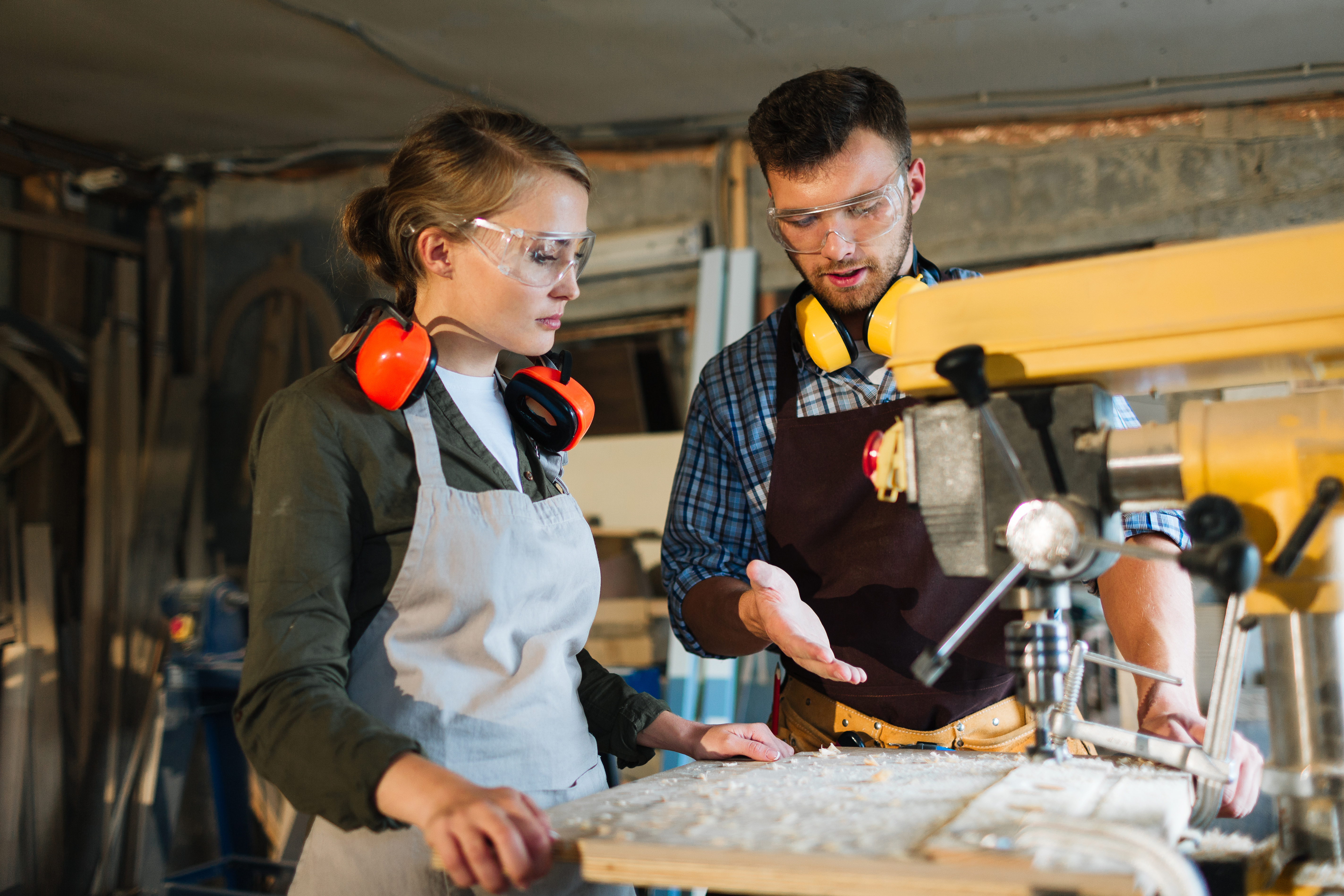 A man is explaining to a woman how to use woodworking equipment. They are both wearing protective gear.