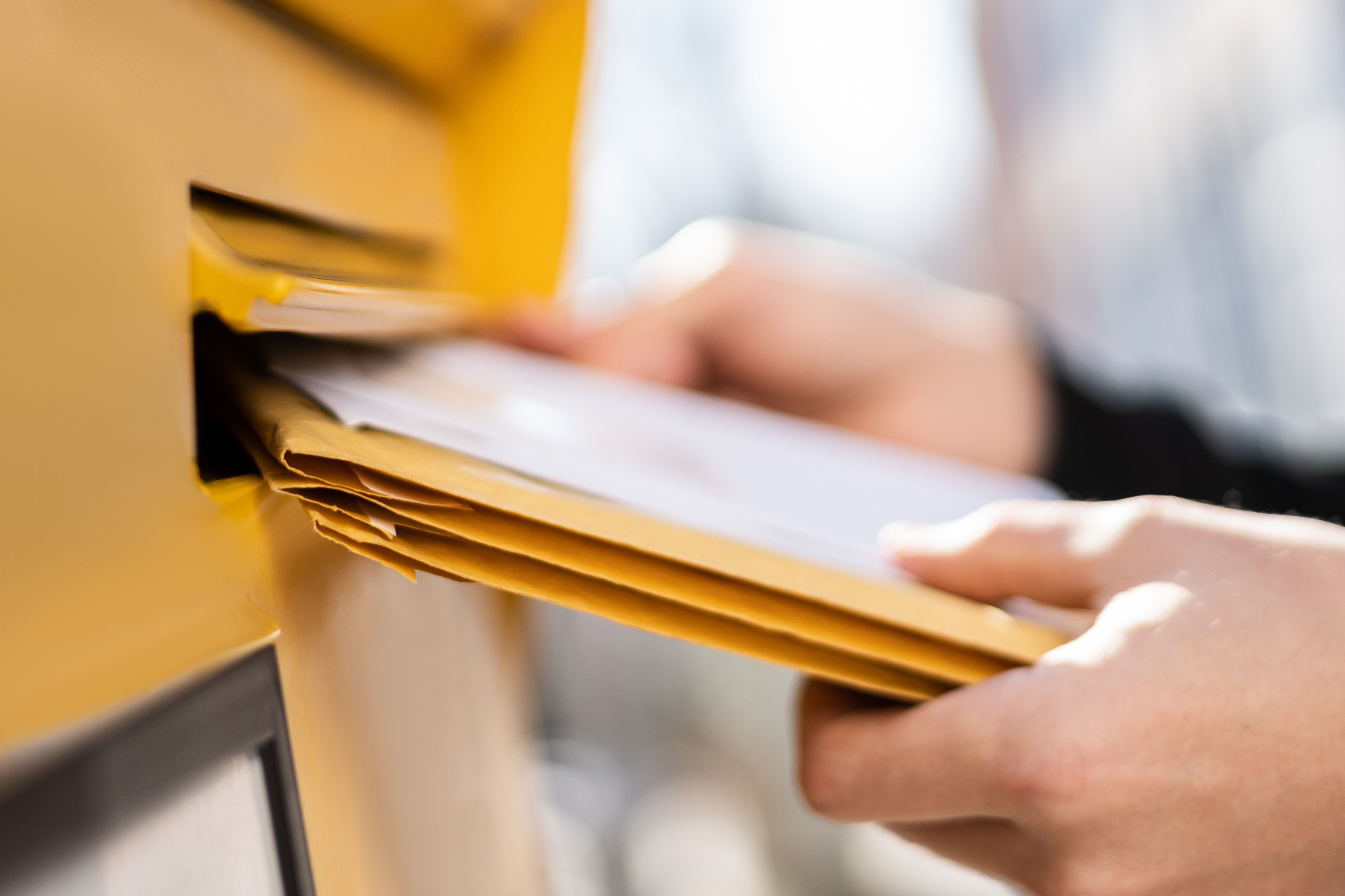 Image shows a person's hands putting yellow envelopes into a mailbox slot.
