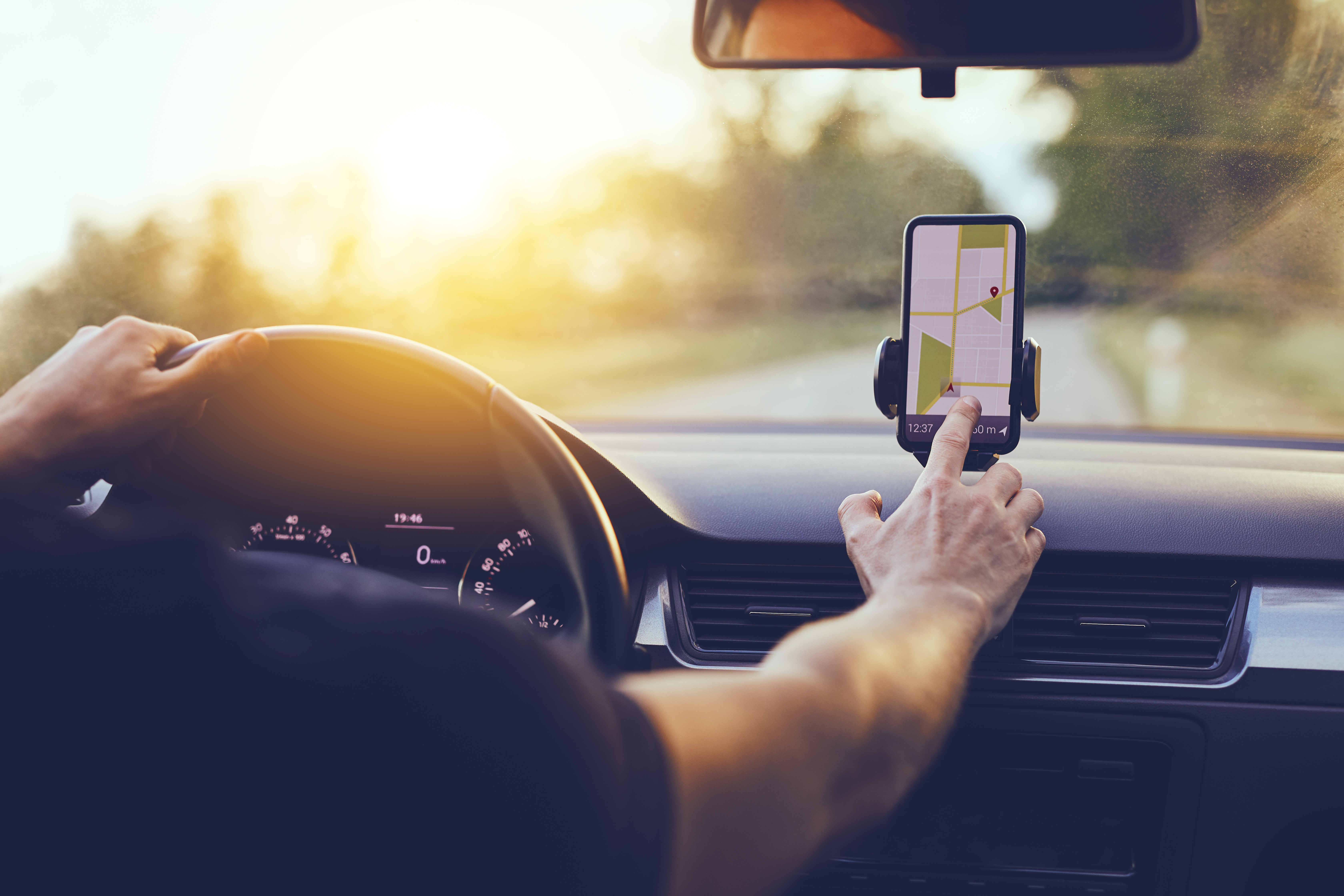 The image shows the arms of a man sitting in the driver's seat of a car. His left hand is resting on the steering wheel, while the right is using the GPS on his phone to navigate.