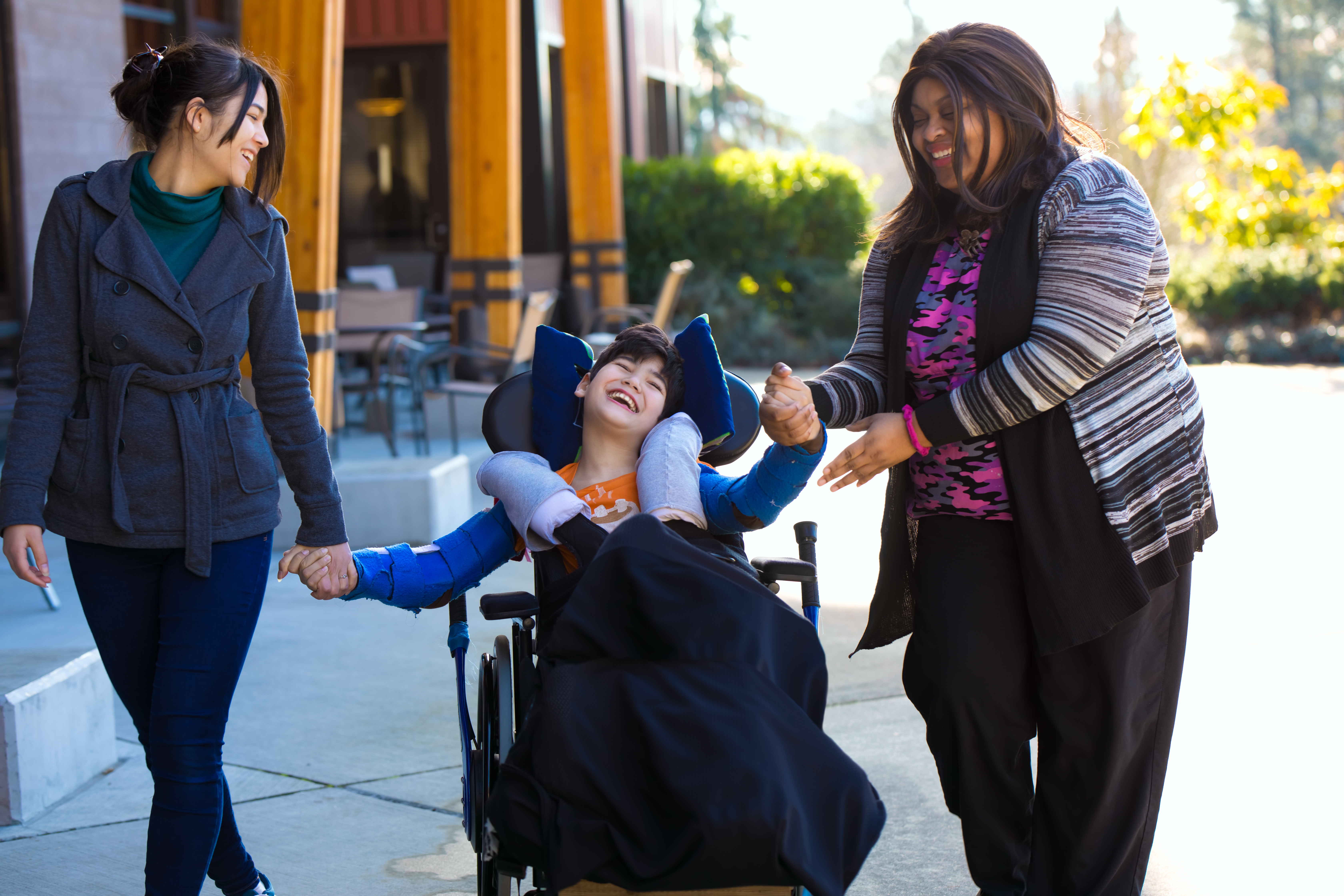 Image shows a male student with special needs in a wheelchair accompanied by two adult women on either side. The women are each holding one of his hands and looking at the boy. They are all smiling.