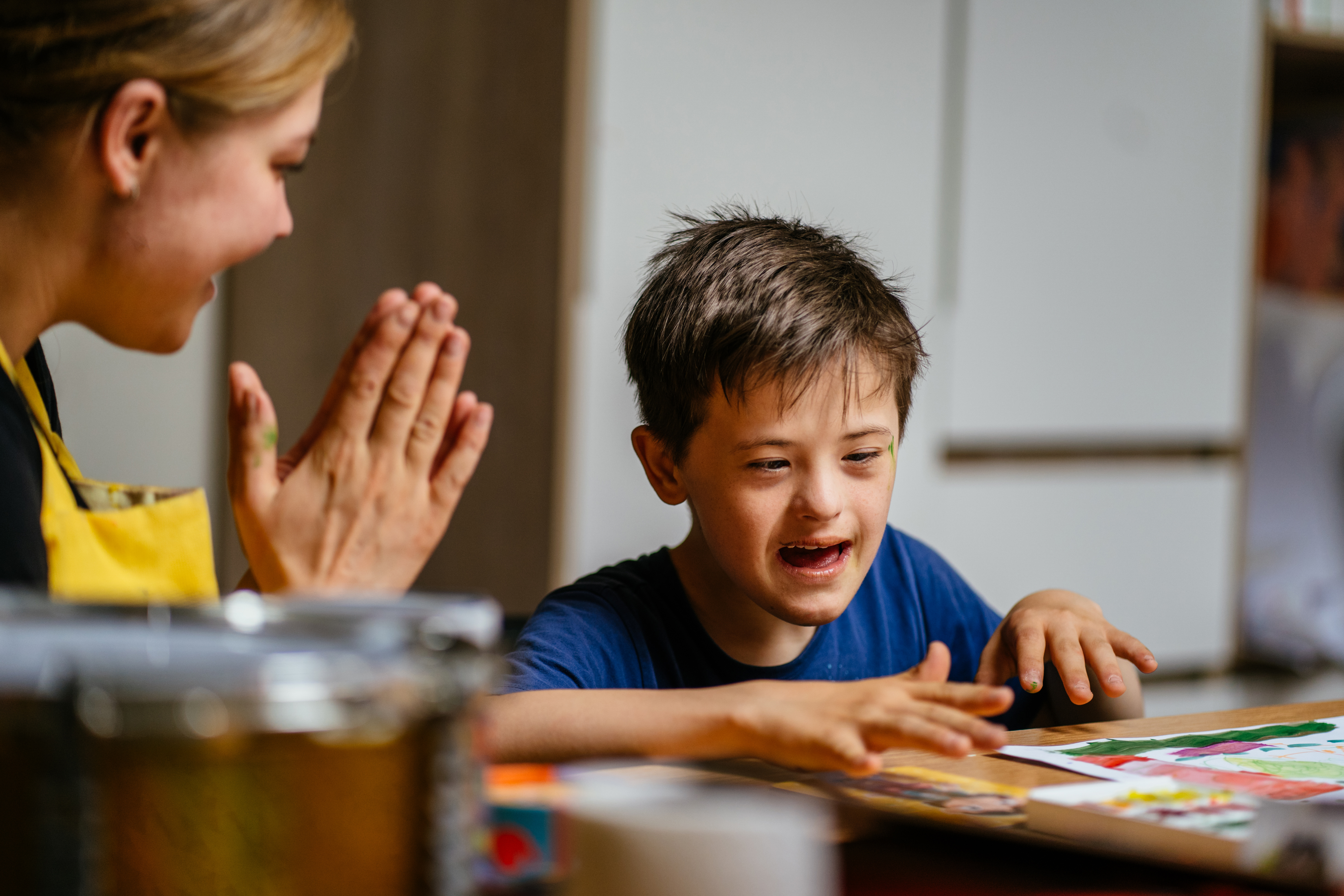 Image shows a boy with special needs doing an art project at a desk. A female teacher beside him is clapping and they both look happy.