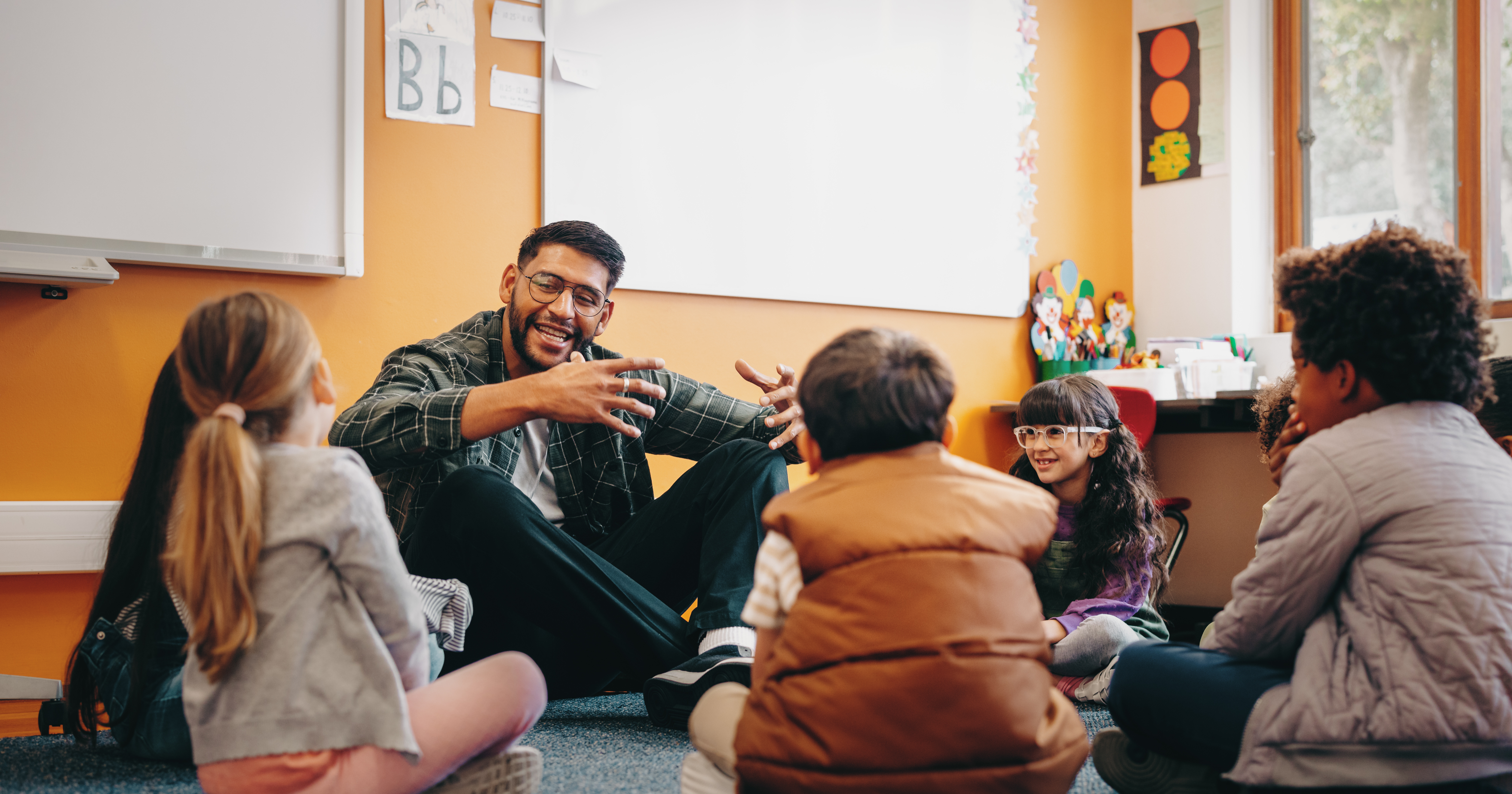 Image shows a teacher and a small group of students sitting on the classroom floor in a circle. He is smiling and gesturing and looking at one of the students and all the students are listening attentively to him.