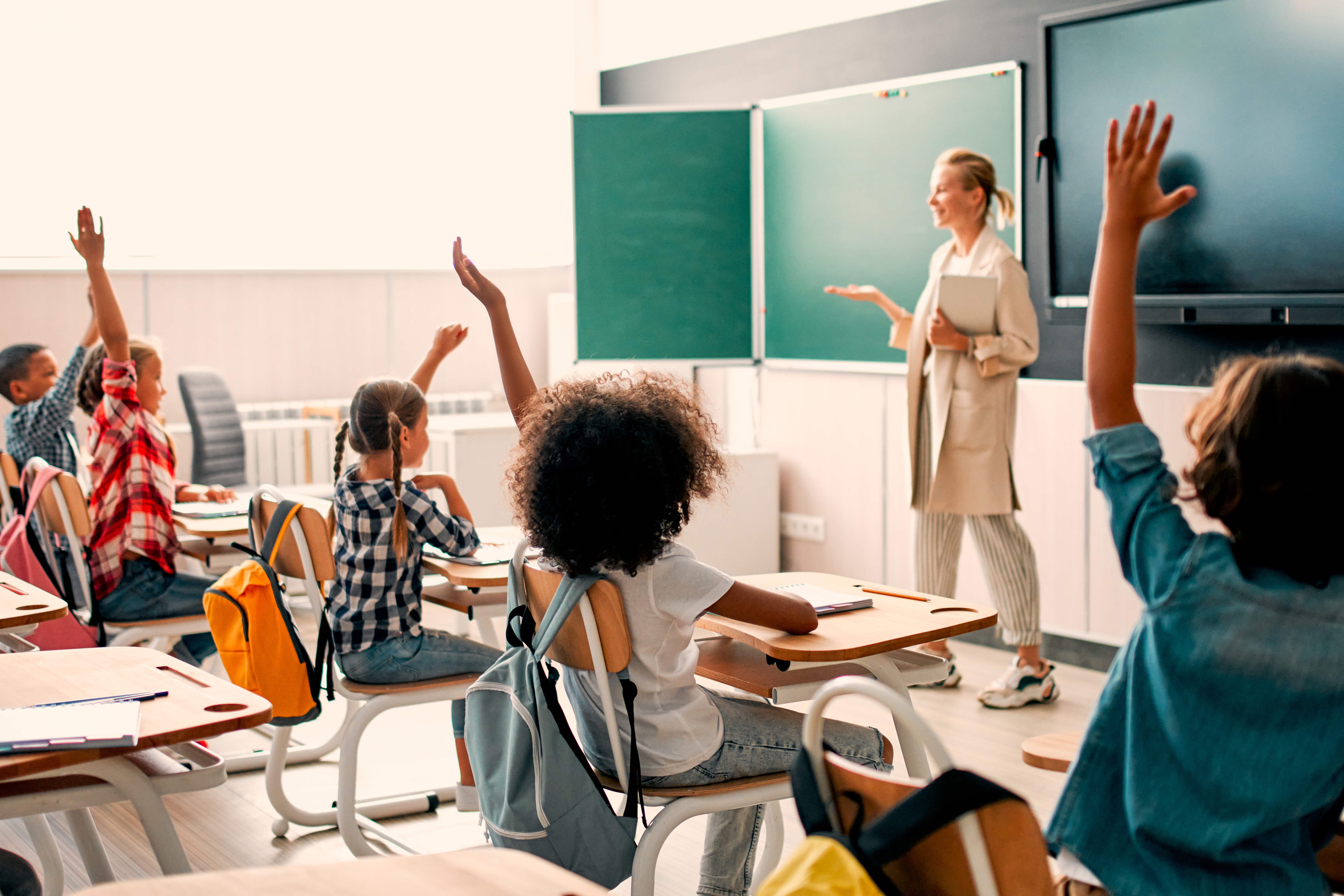 A teacher is standing at the front of a classroom and has her hand out as if she has asked a question and is waiting for someone to volunteer to answer. Students are looking at her with their hands raised to be called on.