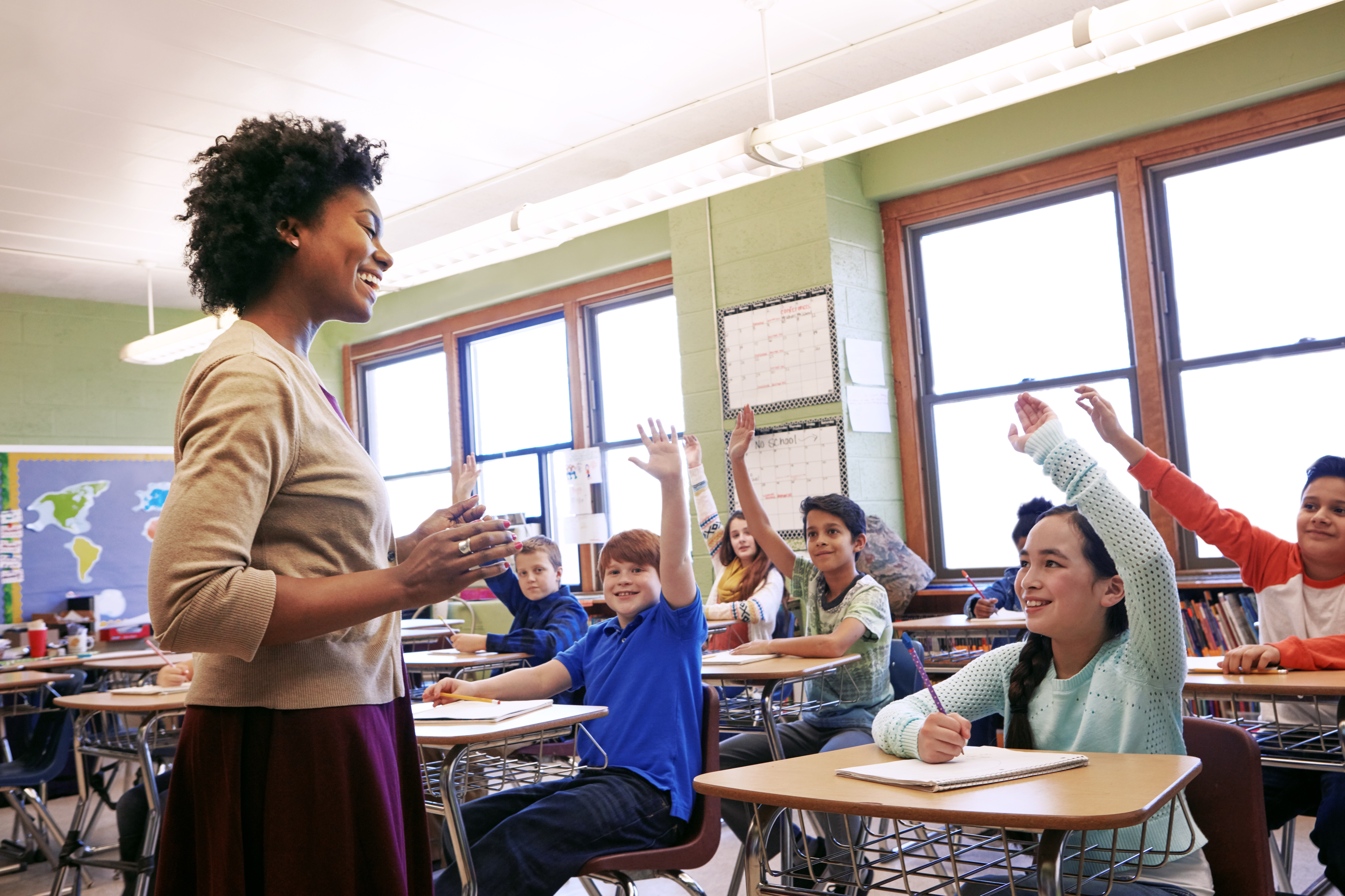Image shows a female teacher in the foreground smiling and looking at her students who are all sitting at their desks. Each student has their hand raised and is looking excitedly at their teacher.