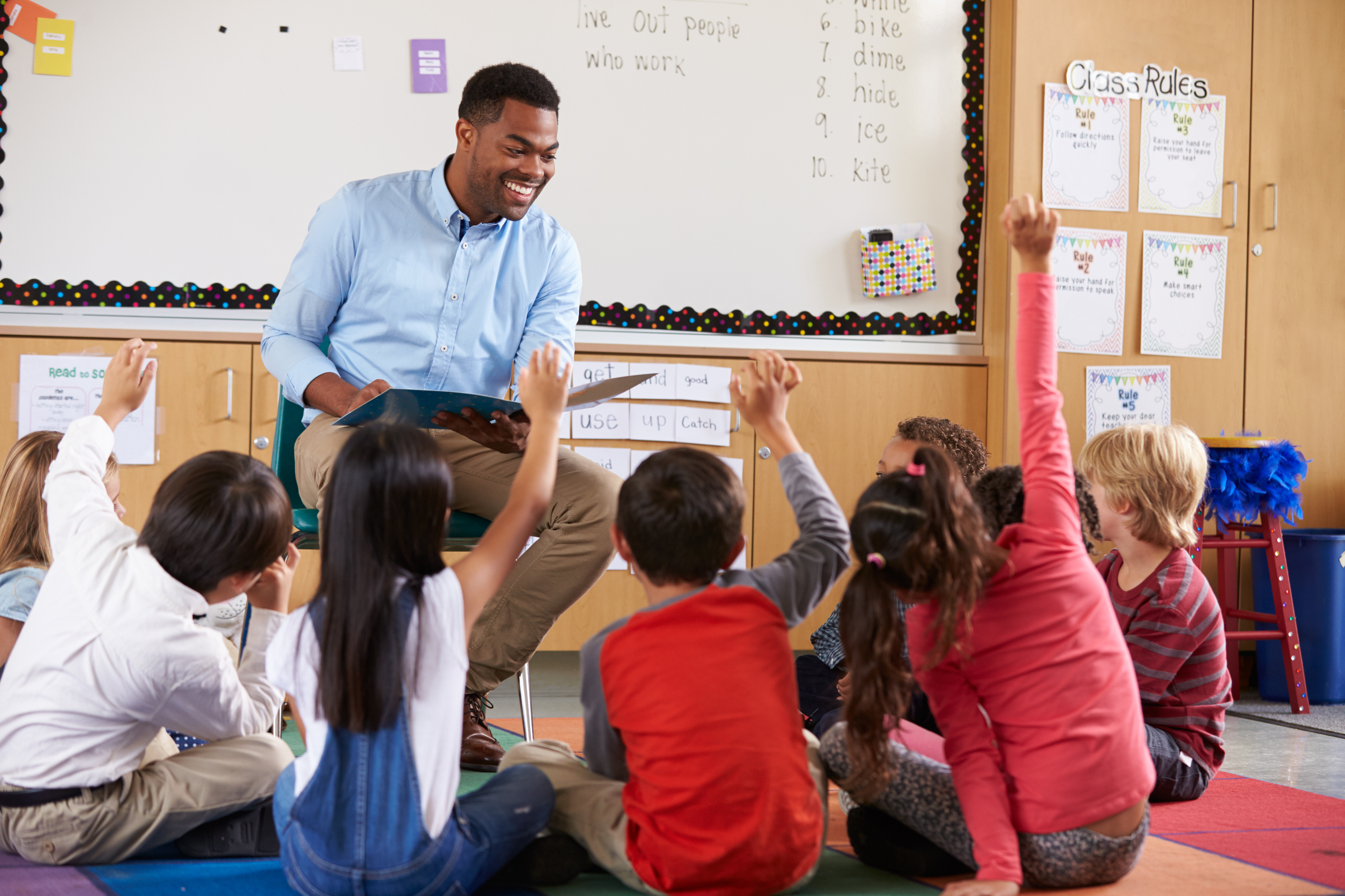 A male teacher is sitting on a chair in a classroom reading a book to a group of students sitting on the floor. He is smiling and some of the students have their hands raised.