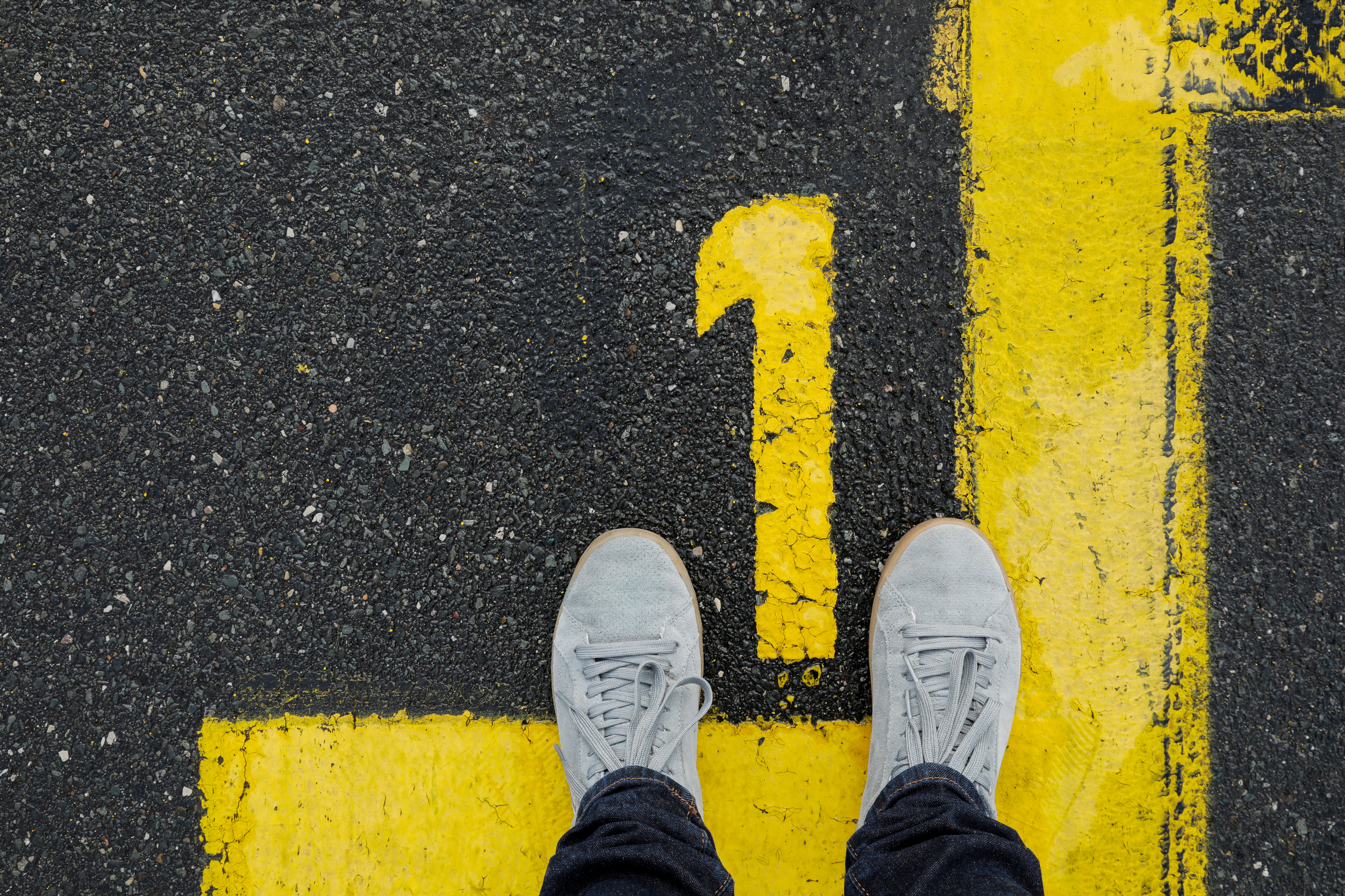 Image shows a yellow number 1 painted on an asphalt road, with a person's feet in sneakers pointed toward it.