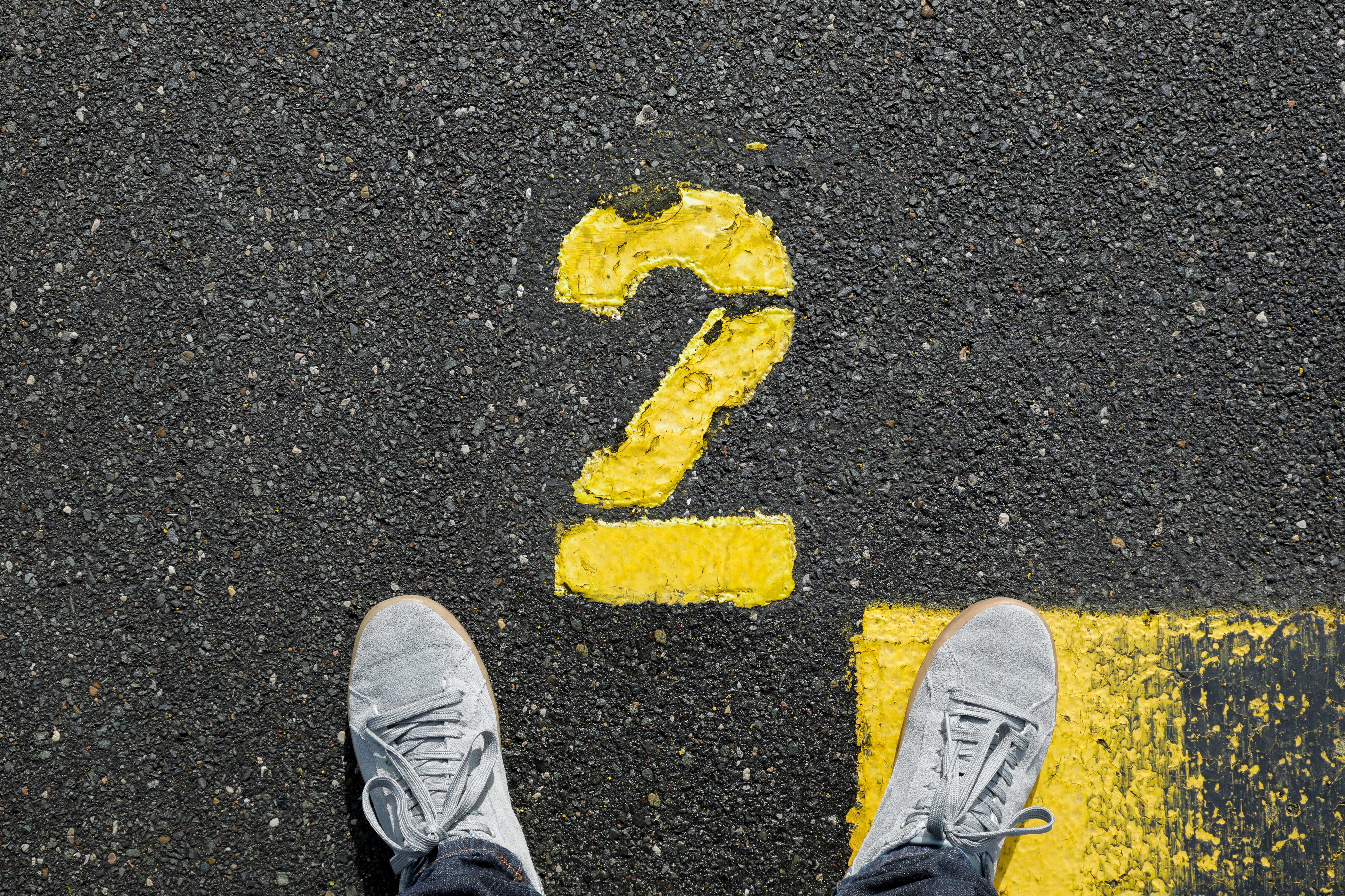 Image shows a yellow number 2 painted on an asphalt road, with a person's feet in sneakers pointed toward it.