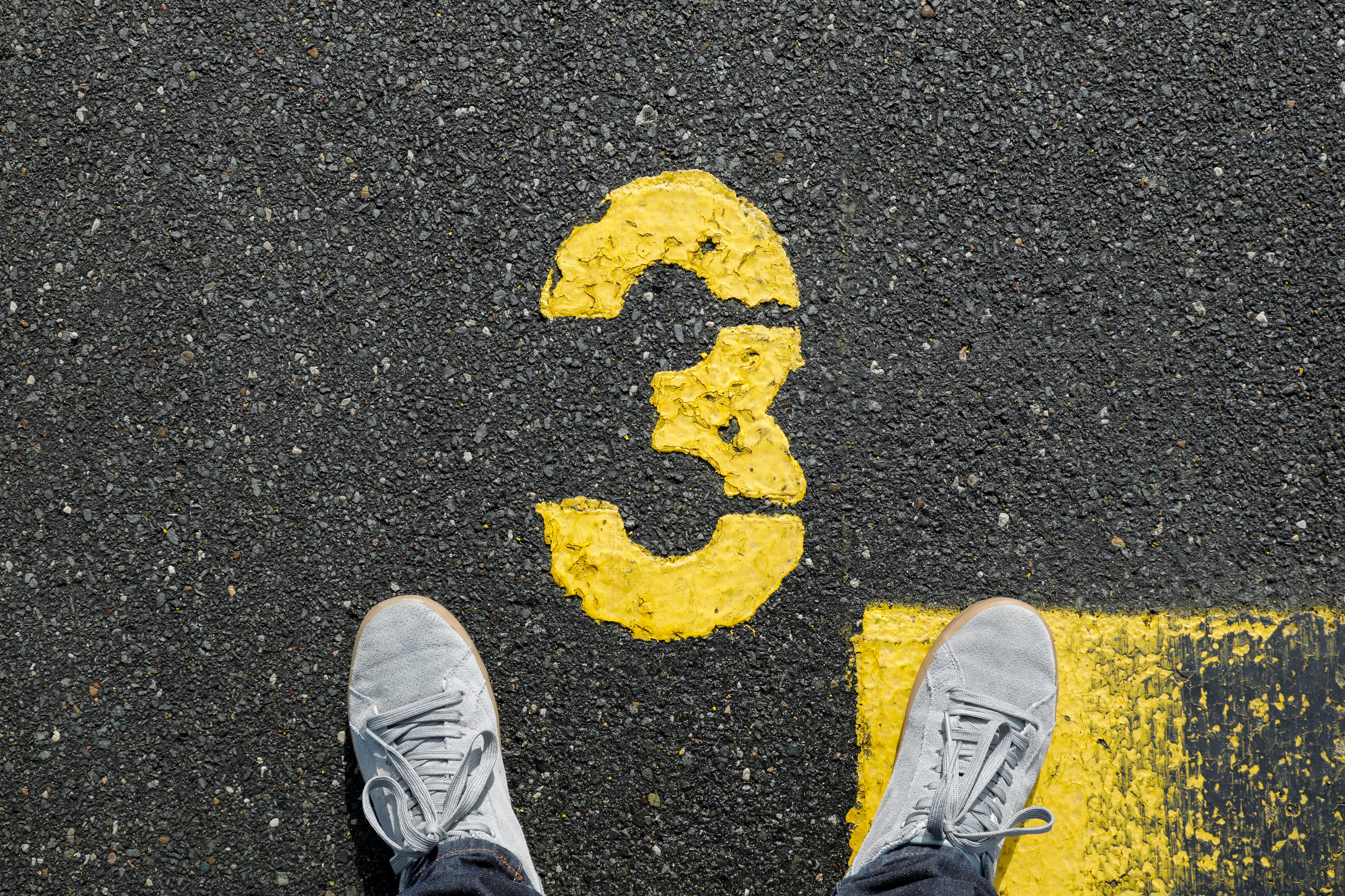 Image shows a yellow number 3 painted on an asphalt road, with a person's feet in sneakers pointed toward it.