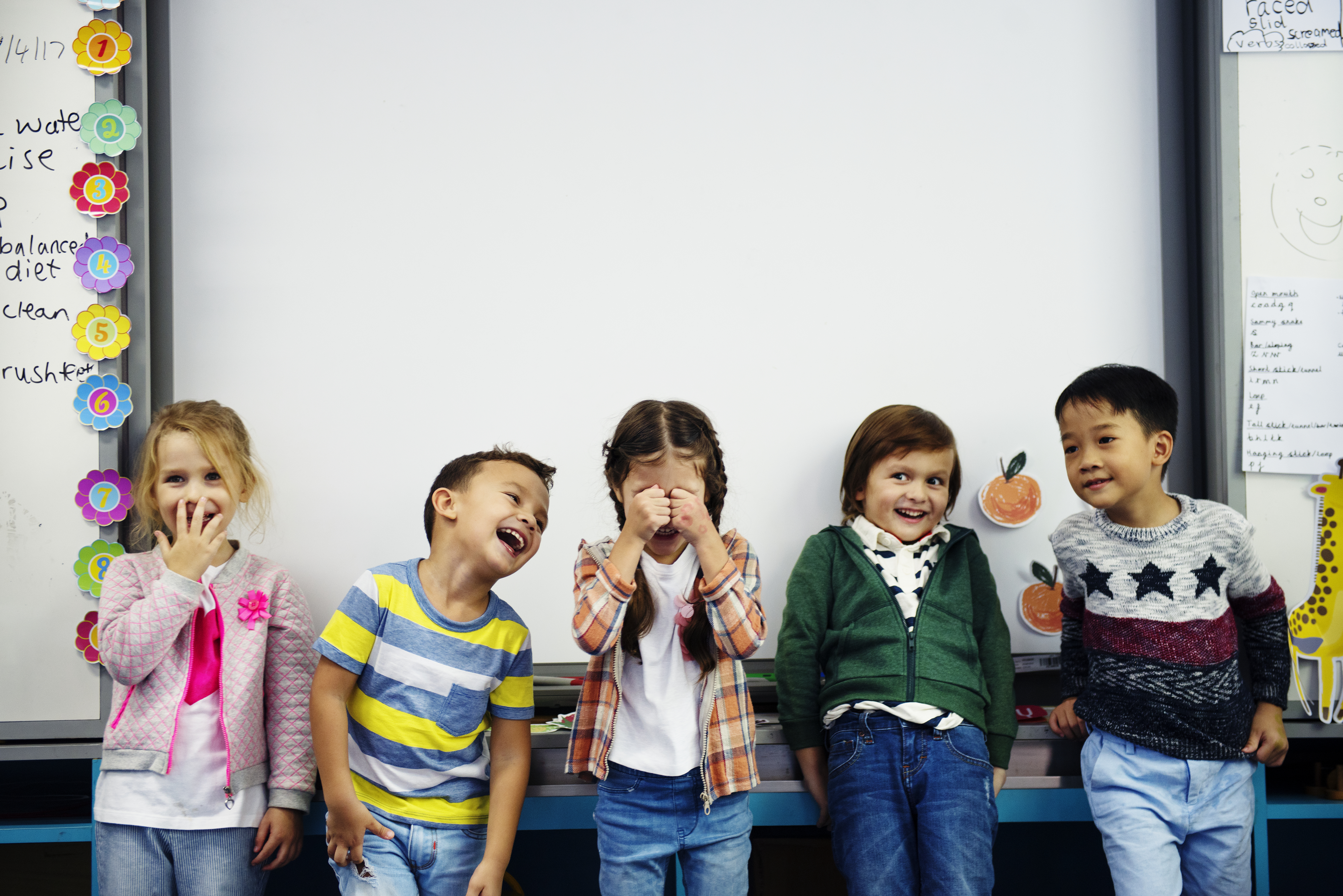 Five young students in the PK-3 age group are standing in front of a whiteboard with different happy expressions. Image by rawpixel.com on Freepik